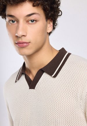 Jonge man met krullend haar die een beige geperforeerd gebreid shirt met een donkerbruine kraag draagt, en lichtjes opzij kijkt.