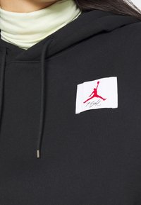 Zwarte hoodie met een wit label met een rood Jumpman-logo. De trekkoorden zijn metallic; de zachte stoftextuur is zichtbaar. Gelaagd met een lichtgroene coltrui.