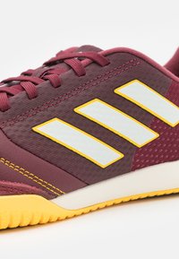 Scarpa da ginnastica color bordeaux con materiale testurizzato, tre strisce bianche bordate di giallo e suola in gomma gialla. Presenta un design con lacci.