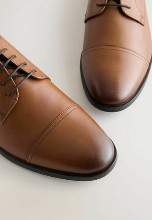 Een paar gepolijste bruine leren dresschoenen met zwarte zolen en donkerbruine veters op een lichte achtergrond.