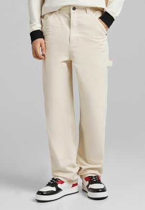 Personne portant un pantalon beige à jambes droites avec des poches et des baskets blanches, noires et rouges, debout devant un fond uni.