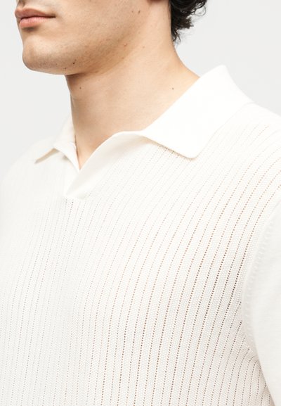 Κοντινό πλάνο ενός ατόμου που φοράει λευκό ριμπ πουλόβερ πόλο με διασκορπισμένο γιακά σε απλό ανοιχτό φόντο.