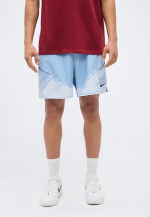 Személy világoskék Nike rövidnadrágot visel fehér absztrakt mintával, piros pólót, fehér zoknit és fehér Nike tornacipőt, egyszínű háttér előtt állva.
