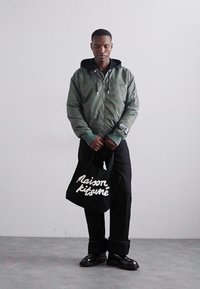 Πράσινο bomber jacket, μαύρη κουκούλα, μαύρο παντελόνι και μαύρα παπούτσια. Λευκή τσάντα tote με κείμενο "Maison kitsuné". Απλός σχεδιασμός, χαλαρή εφαρμογή.