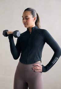 Veste de sport noire à zip avec manches longues, associée à des leggings marron taille haute. La personne tient un haltère noir hexagonal.