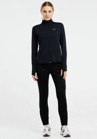 Marineblaue langärmlige Jacke mit Reißverschluss, Stehkragen, schwarze Leggings und weiße Sneakers mit bunten Akzenten. Glatter Stoff und taillierte Passform.