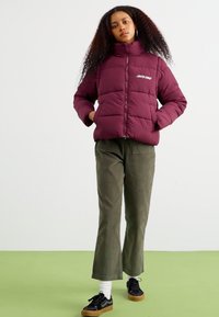 Burgundy puffer jacket with horizontal padding, high collar, and front zipper; paired with olive green wide-leg pants and black sneakers.