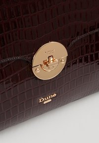 Burgundy patenterad läderhandväska med krokodilmönster, med en guldfärgad metallspänne och präglad logotyp på framsidan.