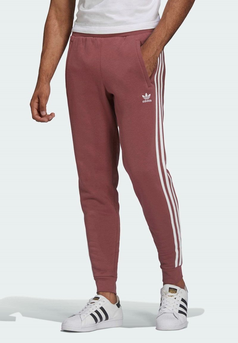 Vínové teplákové kalhoty s bílým třípruhovým designem, bočními kapsami a žebrovanými lemy. Noste s bílými teniskami Adidas.