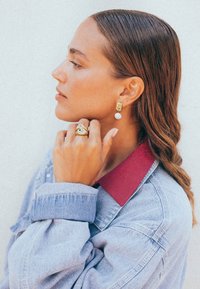 Guldörhängen med färgglada accenter och en vit pärla, kombinerade med en guldring med blå stenar. Bärs med en jeansjacka och röd krage.