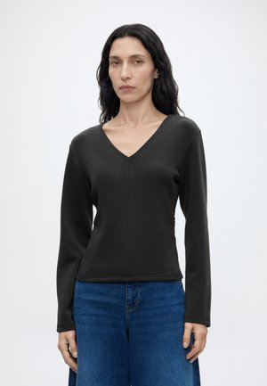 Femme aux longs cheveux foncés portant un haut noir à col en V à manches longues et un jean bleu, debout devant un fond clair uni.