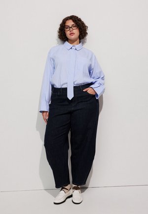 Frau mit lockigem Haar und Brille, die ein hellblaues übergroßes Hemd, lockere dunkle Hose und weiße Schuhe trägt und an einer weißen Wand steht.