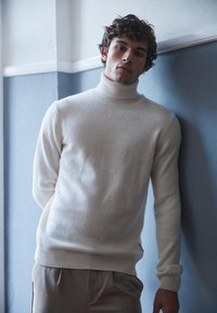 Romige coltrui gemaakt van een zacht materiaal, met ribbels aan de mouwen en zoom. G gedragen met beige broeken, tegen een blauwe muur.