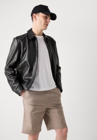 Sort læderjakke med en glat finish, hvid t-shirt under og beige shorts. Modellen bærer en sort cap, hænderne ilommerne.
