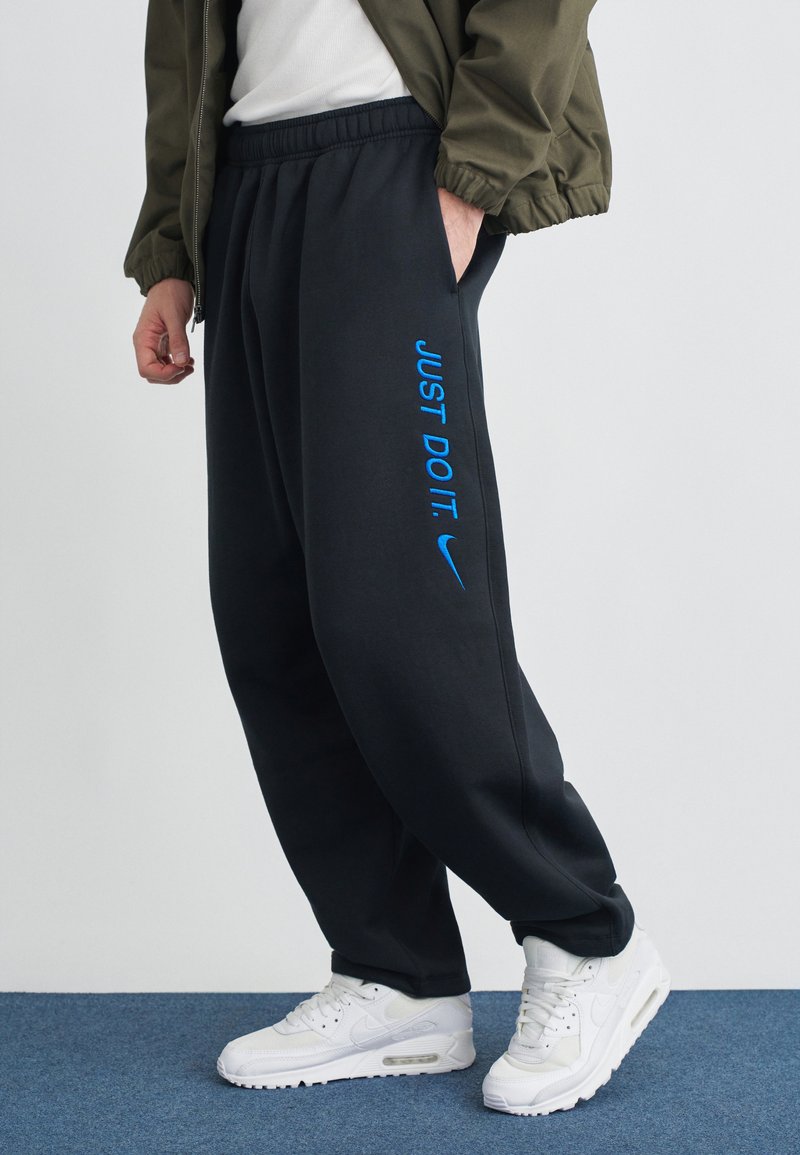 Melnas sporta bikses ar elastīgu jostu, uz kurām ir zilā krāsā uzraksts "JUST DO IT." un Nike logotips; valkātas ar baltām sporta kurpēm uz zila fona.