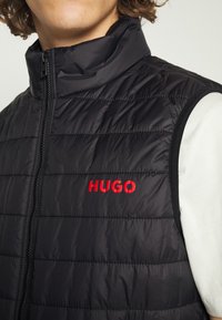 Gilet imbottito nero con alto colletto, caratterizzato da cuciture orizzontali e un prominente logo rosso "HUGO" sul petto. Chiusura frontale con zip.