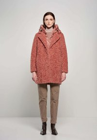 Różowy sztuczny futrzany płaszcz z dużym kołnierzem, nałożony na beżowy wzorzysty top, w połączeniu z beżowymi spodniami z cordurowego materiału i brązowymi botkami za kostkę.