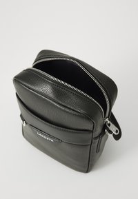 Sort tekstureret Lacoste crossbody taske med frontlomme, sølvlynlås delvist åben og mærkelogo på frontlommen.