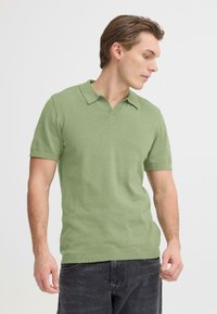 Polo à manches courtes en coton vert clair, avec un col en V, des bords nets et une texture douce. Porté avec un jean en denim foncé.