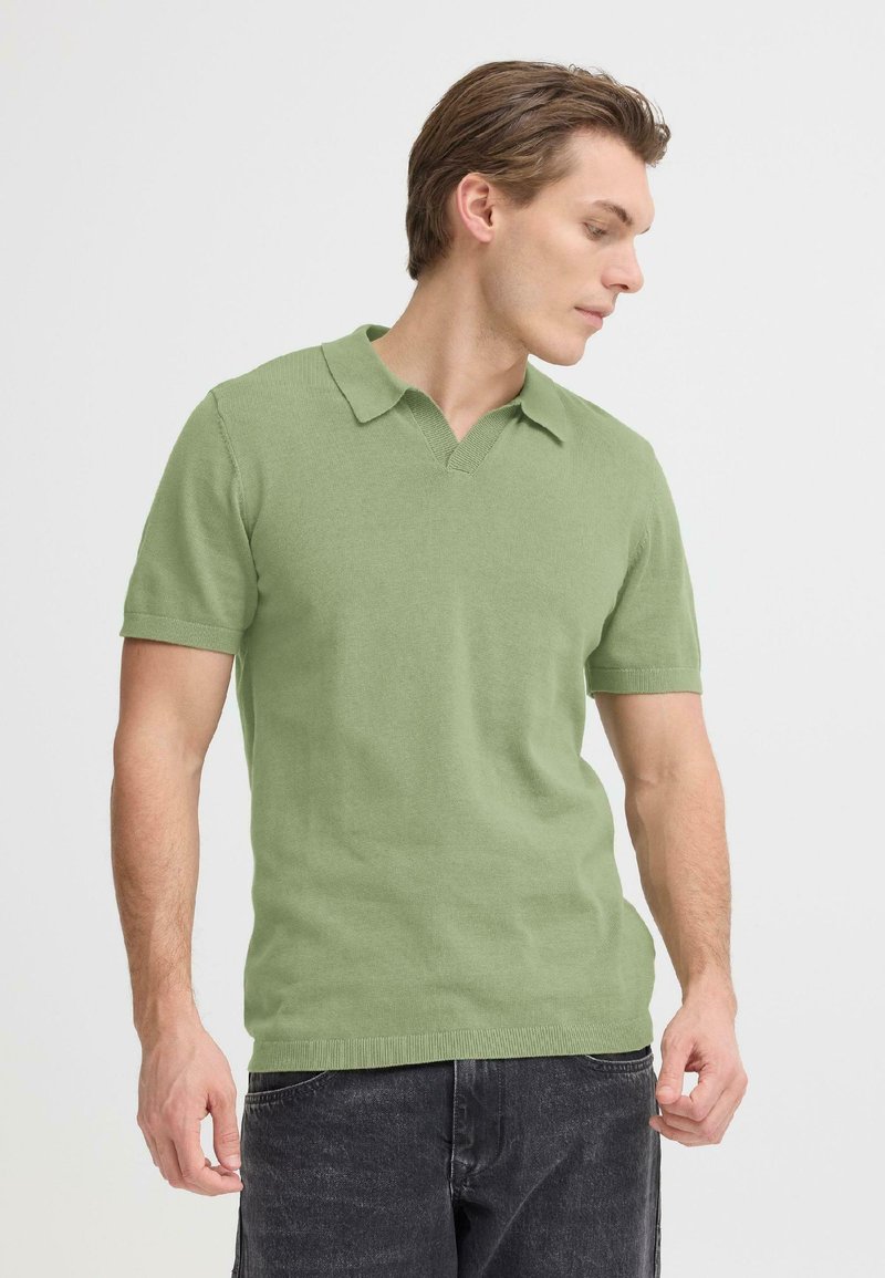 Polo à manches courtes en coton vert clair, avec un col en V, des bords nets et une texture douce. Porté avec un jean en denim foncé.