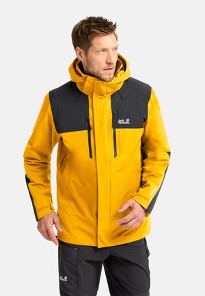Man in een geel-zwarte outdoorjas en zwarte broek kijkt naar de zijkant, staand tegen een effen lichte achtergrond.