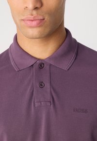 Fialové polo tričko s texturovanou tkaninou, dvěma knoflíky a malým embosovaným logem na levé straně. Klasický design límce.