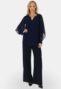 Granatowa bluzka z lekkimi, przezroczystymi rękawami, z dekoltem w kształcie łezki i srebrnym akcentem. Dopasowana do szerokich granatowych spodni.