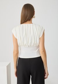 Biała bluzka z krótkimi rękawami, strukturalne fałdy z tyłu, dopasowany krój, w połączeniu z czarnymi spodniami. Gładki materiał, minimalistyczny design.