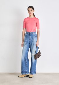 Top cor-de-rosa de mangas curtas, jeans de cintura alta azul claro com detalhes desgastados e botas amarelas de cano curto. Bolsa marrom de ombro completa o visual.