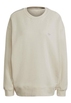 Person trägt beige Adidas-Sweatshirt mit geprägtem Logo und schwarze Hose mit weißen Streifen, blickt nach hinten mit leicht ausgestreckten Armen.