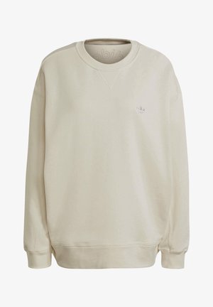 Person trägt beige Adidas-Sweatshirt mit geprägtem Logo und schwarze Hose mit weißen Streifen, blickt nach hinten mit leicht ausgestreckten Armen.