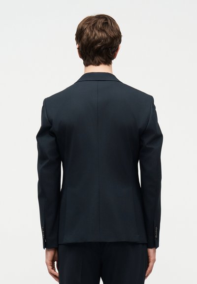 Άνδρας με κοντά καστανά μαλλιά, ντυμένος με εφαρμοστό σκούρο μπλε σακάκι κοστουμιού και παντελόνι, στέκεται γυρισμένος πλάτη σε απλό λευκό φόντο.