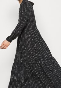 Černé maxi šaty s dlouhými rukávy, které mají bílý tečkovaný vzor, splývavý vrstevnatý design a kulatý výstřih.