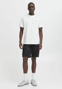 Camiseta de algodón blanca con el texto "BUENA VIDA", combinada con pantalones cortos cargo negros con bolsillos laterales y zapatillas grises con calcetines blancos.