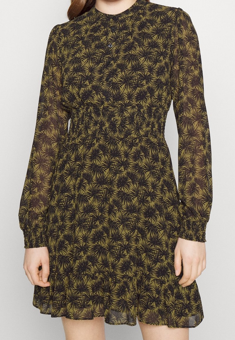 Femme portant une robe longue à manches longues à motif floral vert et noir, avec une taille froncée et un col boutonné, debout sur un fond uni.