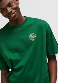 Grön bomullströja med rund halsringning och korta ärmar. Den har en liten gul logotyp med lagerblad på bröstet.