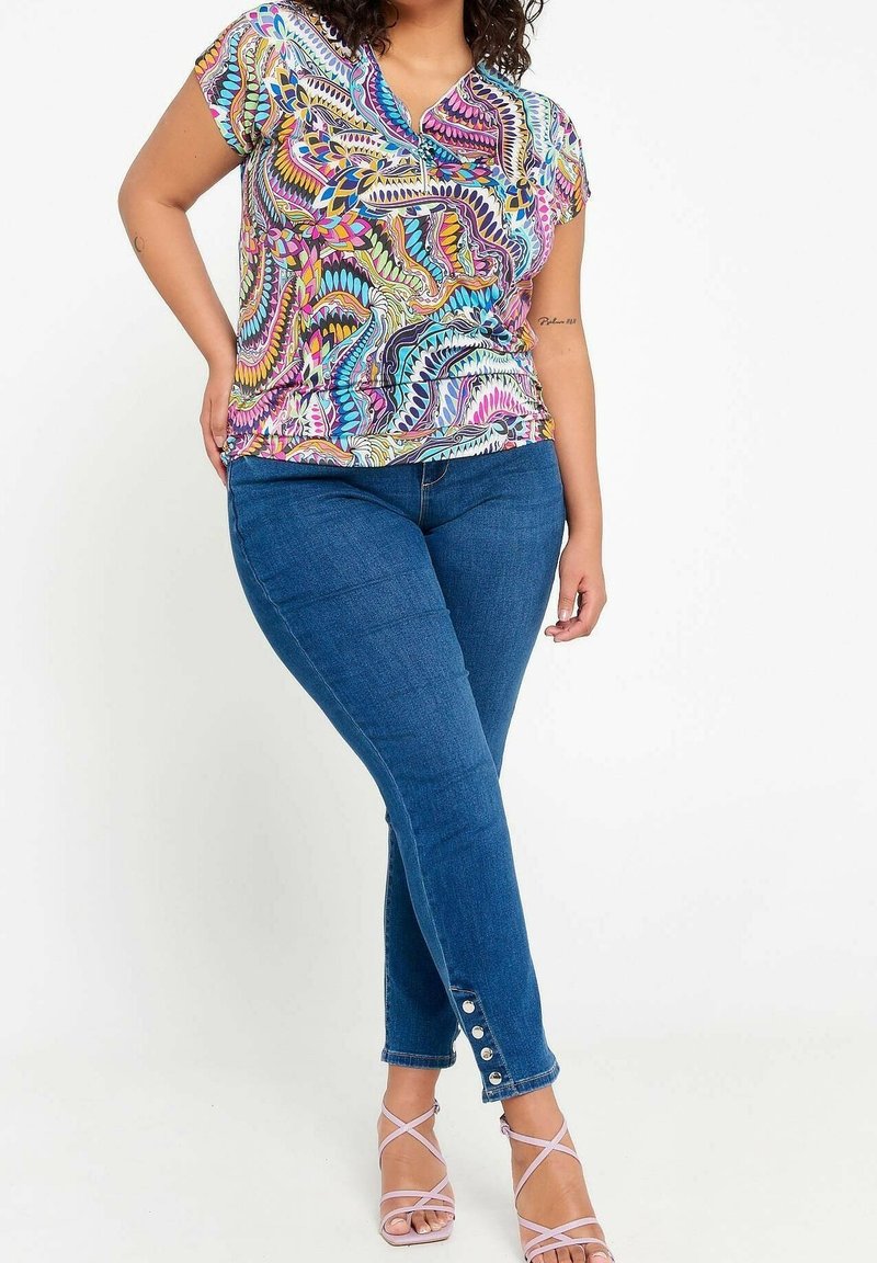 Donna in una maglietta a maniche corte con un motivo colorato, jeans blu aderenti con dettagli a bottoni alle caviglie e sandali con tacco alto e strisce rosa.