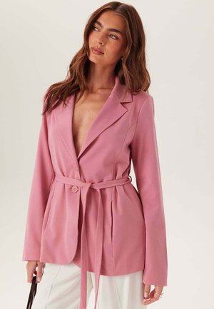 Kvinde med langt brunt hår iført en pink blazer med bælte og hvide bukser, der holder en sort håndtaske og ser let mod venstre.