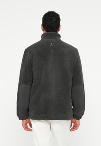 Vīrietis ar muguru pret kameru, ģērbies tumši pelēkā flīsa jakā ar pastiprinātām elkoņu zonām un baltām biksēm, uz viendabīga fona.