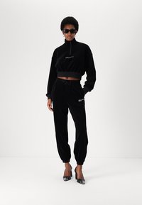 Black velvet tracksuit with cropped zip-up top and tapered pants. Features elastic waistband, logo detail, and paired with shiny black heels.