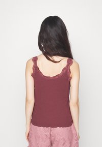 Donna con lunghi capelli scuri indossa un top senza maniche color borgogna con bordi in pizzo e pantaloni corti di pizzo rosa, in piedi di fronte a uno sfondo bianco.