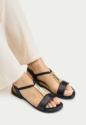 Stopy w czarnych płaskich sandałach z złotymi kwadratowymi ozdobami, zestawione z jasnobeżowymi spodniami na białym tle.