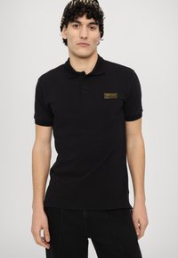Črna polo majica s slim fit krojem, ki ima ovratnik s tremi gumbi in zlate barve blagovno znamko na levem prsnem delu. Oblečena z črnimi kavbojkami.