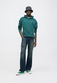 Zielona bluza z kapturem, niebieskie dżinsy, czarna czapka typu bucket, zielone sneakersy. Swobodny strój z przednią kieszenią na bluzie i prostymi dżinsami.