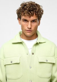 Lichtgroene fleecjas met een kraag, drukknoopsluiting en twee voorzakken met zilveren accenten. Glad textuur.