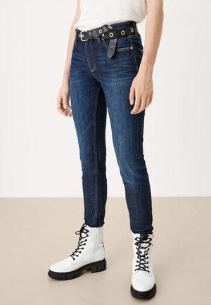 Person iført mørkeblå tætsiddende jeans, hvide snørestøvler, sort bælte med store sølvnitter og en hvid top puttet ned i bukserne.