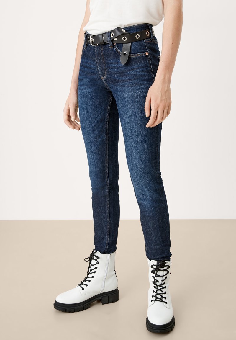Personne portant un jean skinny bleu marine, des bottes de combat blanches à lacets, une ceinture noire avec de grands œillets argentés et un haut blanc rentré.