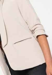 Jasno beżowa marynarka o gładkiej fakturze, z wyłogami i podsumowanymi rękawami. Posiada boczną kieszeń oraz dopasowany, skrojony fason.