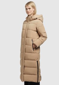 Lang beige pufferjakke med hætte, der har vandrette quiltede sektioner, sidelommer med lynlås og lige kant. Glat, mat stoftekstur.