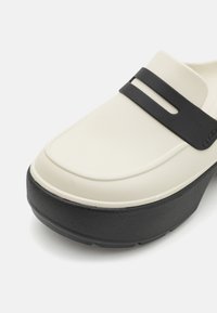 Krämfärgad slip-on sko med en strukturerad yta, som har ett svart gummiband och en tjock svart plattformssula.